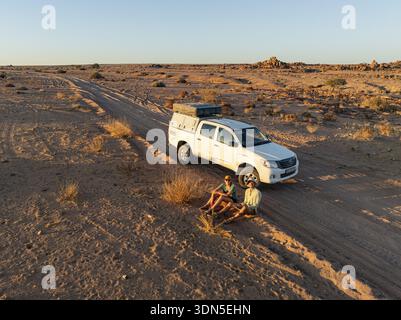 Zwei Reisende sitzen neben einem Geländewagen auf einer Feldstraße in der Wüste, Erongo, Damaraland, Namibia Stockfoto