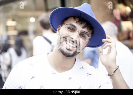 Überschwängliche Cowboy-Mentalität, glücklicher heller Mann mit blauem Hut und Brille, lächelnd in einer hellen, lässigen Umgebung im Freien, Zahnlächeln, Kopfbedeckung, Kopfbedeckung Stockfoto