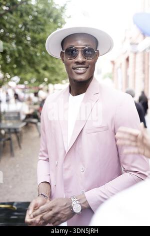 Sanguines Lächeln eines sympathischen, netten Mannes, der mit seinem Kollegen auf der Straße spricht, mit einem stilvollen pinken Blazer und einem weißen Hut, der draußen mit einer Sonnenbrille lächelt Stockfoto