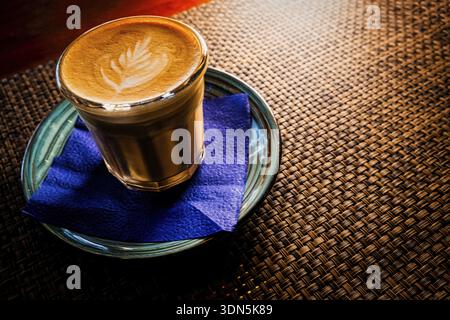Flacher weißer Kaffee wird auf einer Untertasse über dem Tisch mit Bambusmatte mit Hintergrundbeleuchtung serviert, Copyspace Stockfoto