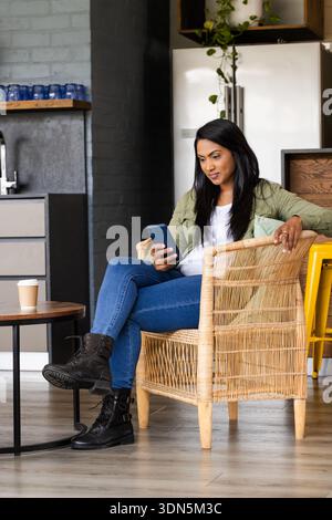 MittelErwachsene indische Frau, die auf Korbstuhl in der Küche sitzt, Smartphone hält, Kaffeetasse ruht Stockfoto