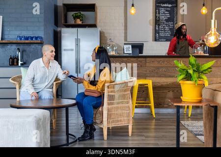 Verschiedene Freundinnen unterhalten sich und nutzen ihr Smartphone in der Lounge des Coffee Shops mit Korbsesseln Stockfoto