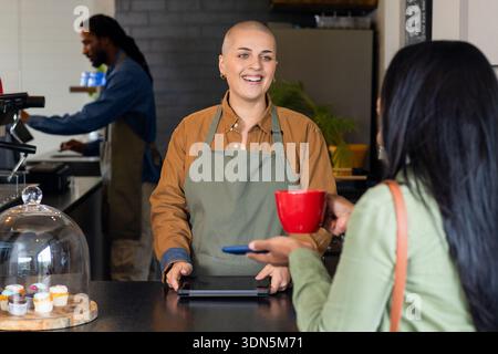 Lächelnde Frau Barista, die ein Tablet anzapft, um einer indischen Kundin Kaffee an der Theke zu servieren Stockfoto