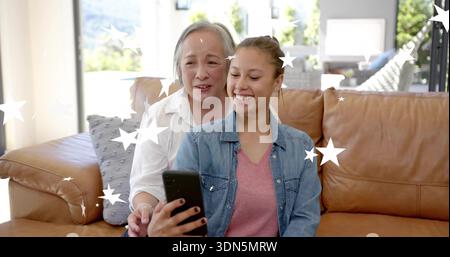 Lächelnde Jeansjacke Teenager hält Smartphone auf Ledersofa, Frau in weißer Bluse lehnt sich hinein Stockfoto