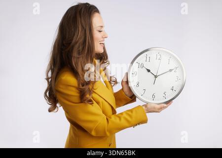 Eine lebendige Geschäftsfrau in gelbem Blazer lächelt auf eine analoge Uhr und symbolisiert die Freude an erfolgreichem Zeitmanagement und Geschäftstreffen Stockfoto