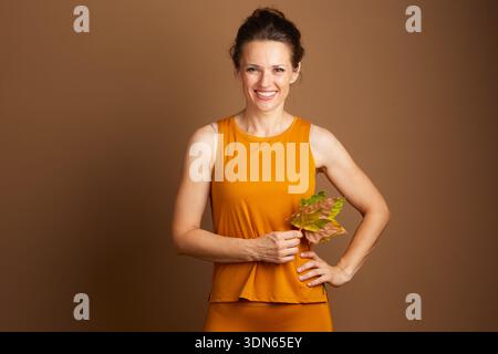 Eine lächelnde junge Frau in senfgelber Aktivkleidung hält einen Haufen bunter Herbstblätter und vermittelt eine warme und gesunde jahreszeitliche Stimmung gegen ein Uni Stockfoto