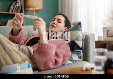 Eine Frau liegt auf einem gemütlichen Sofa, in eine Decke gewickelt, und liest aufmerksam ein Thermometer. Die ruhige, sanfte Beleuchtung und die heimelige Atmosphäre spiegeln einen Moment von wider Stockfoto