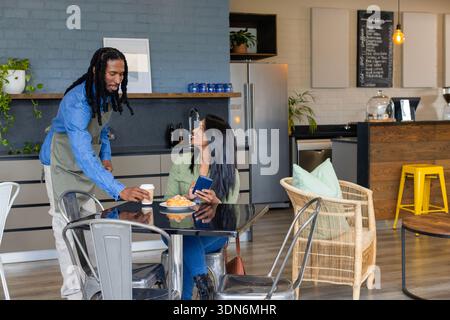 Afroamerikanischer Server in grüner Schürze, der asiatische Frau, die ihr Smartphone im Café hält, eine Kaffeetasse überreicht Stockfoto
