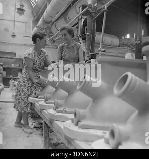 Slowiansk, Region Donezk, Ukraine, UdSSR. 1960er Jahre Historisches Archivfoto. Zwei weibliche Arbeiter untersuchen komplexe Keramikverteiler am Sloviansk Ceramic Mähdrescher. Die jüngere Frau in einem gemusterten Sommerkleid untersucht eine schwere Tonkomponente mit mehreren Ausgängen. Ihr erfahrener Kollege steht neben ihr und überwacht den Prozess. Der Fabrikboden ist mit identischen säurebeständigen Industrieteilen ausgekleidet. Lüftungsrohre sind sichtbar. Dieses authentische Schwarz-weiß-Bild fängt die Mentorschaft und die tägliche Arbeit sowjetischer Frauen im friedlichen Donbass ein Stockfoto