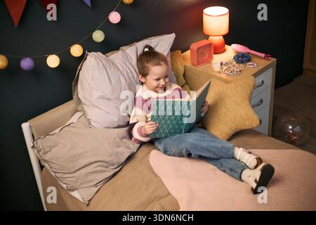 Kaukasisches Mädchen sitzt auf dem Bett und liest Buch mit Lächeln, hält Buch offen in den Händen, umgeben von gemütlichem Schlafzimmer Dekor, Lampe sichtbar auf Nachttisch, sieht verlobt aus Stockfoto