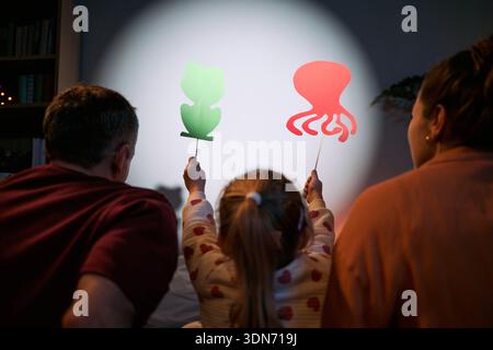 Kaukasischer Mann mittleren Alters, kaukasische Frau mittleren Alters und kaukasisches Kind sitzen zusammen, halten tierförmige Schattenpuppen vor der beleuchteten Wand und schaffen verspielte Silhouetten Stockfoto