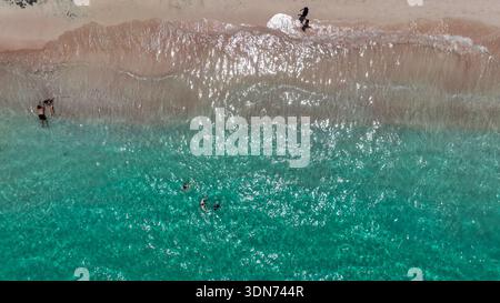 Aus der Vogelperspektive von Pink Beach zeigt korallblaues Wasser, das den sanften, rosa Sand küsst und von Schwimmern im Schatten der Küstenbäume gesäumt ist. Stockfoto