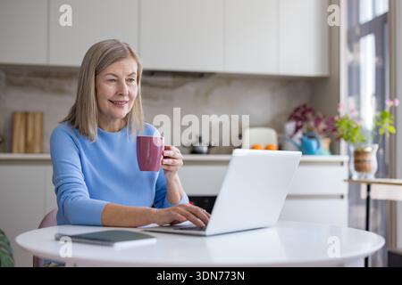 Eine ältere Frau lächelt und tippt auf einem Laptop, hält einen Becher und arbeitet von zu Hause aus an einem weißen Tisch in ihrer modernen Küche Stockfoto