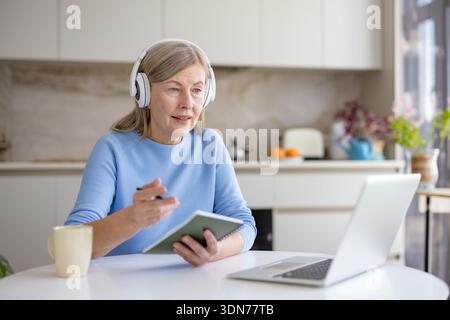 Seniorin, die an einem Videoanruf oder einer E-Learning-Sitzung von ihrer Küche aus teilnimmt, aktiv mit Kopfhörern und einem Notebook teilnimmt und f Stockfoto