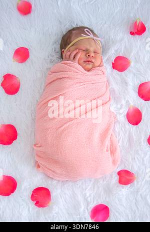Neugeborenes Mädchen, das in rosa Umhüllung schläft, umgeben von Rosenblättern auf weißem Pelz. Stockfoto