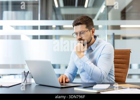 Junger Geschäftsmann, der eine Brille und ein hellblaues Hemd trägt, konzentriert sich tief und denkt, während er an einem Laptop in einer modernen Büroumgebung arbeitet Stockfoto