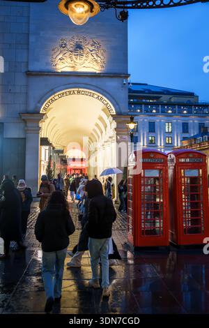 LONDON, GROSSBRITANNIEN. Februar 2025. Leute, die an der Arkade des Royal Opera House und den traditionellen roten Telefonzellen in der Abenddämmerung vorbeilaufen. Stockfoto