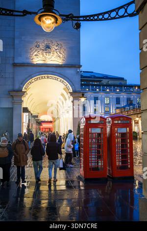 LONDON, GROSSBRITANNIEN. Februar 2025. Leute, die an der Arkade des Royal Opera House und den traditionellen roten Telefonzellen in der Abenddämmerung vorbeilaufen. Stockfoto