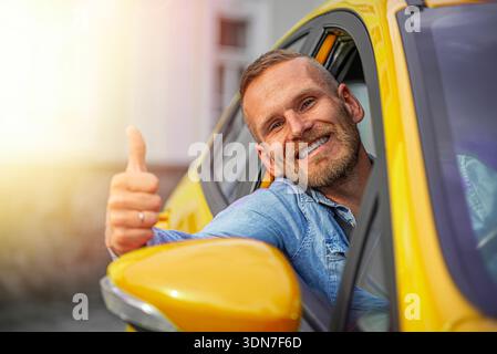 Ein fröhlicher Mann in einem blauen Denim-Hemd beugt sich aus einem hellgelben Autofenster und gibt eine selbstbewusste Daumen-nach-oben-Geste. Stockfoto
