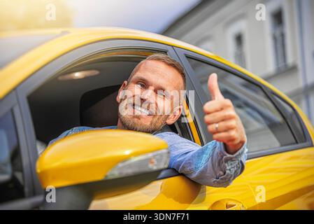 Ein fröhlicher Mann in einem blauen Denim-Hemd beugt sich aus einem hellgelben Autofenster und gibt auf einer sonnigen Stadtstraße die Daumen nach oben. Stockfoto