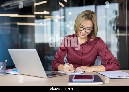 Selbstbewusste Geschäftsfrau mit Brille, die an ihrem modernen Schreibtisch lächelt, Notizen in einem spiralförmigen Notizbuch mit Laptop und Smartphone in der Nähe schreibt, organisiert und konzentriert sich auf die Arbeit Stockfoto