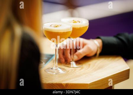 Zwei Orangen-Passionsfrucht-Cocktails ruhen auf einem Holztisch, während die Gäste in der Nähe sitzen. Die Szene zeigt einen entspannten sozialen Moment in einer Bar-Umgebung. Stockfoto