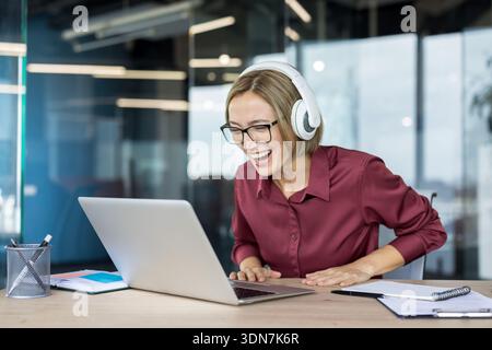 Junge Geschäftsfrau mit Kopfhörern und Brillen lacht aufrichtig, während sie auf ihren Laptop-Bildschirm blickt, positive Nachrichten oder unterhaltsame Inhalte während der Arbeit in einem modernen Büro genießt Stockfoto