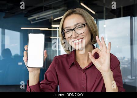 Lächelnde Geschäftsfrau in Brille, die ein Mobiltelefon mit weißem Display präsentiert, während sie eine gute Geste macht, die Erfolg und Anerkennung in einer modernen Büroumgebung vermittelt Stockfoto