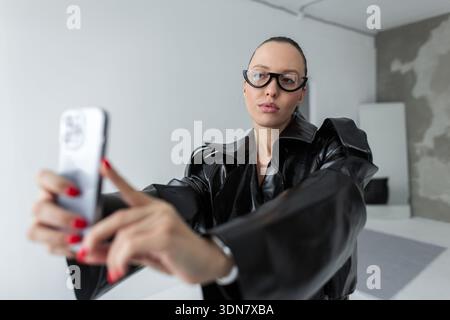 Selbstbewusste, stilvolle Frau in schwarzer Lederjacke und moderner Brille, die ein Selfie mit einem Smartphone in minimalistischer Innenausstattung macht und Mode zeigt, tec Stockfoto