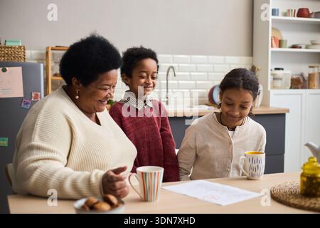 Schwarze Seniorin, die am Küchentisch sitzt, lächelnd und interagierend mit Black Boy und Black Girl, Kinder, die auf dem Tisch zeichnen, während sie Tassen halten, Familienbindung zu Hause Stockfoto