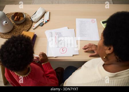 Schwarzes Kind, das neben der schwarzen Seniorin am Tisch sitzt und die Schularbeit mit großer roter F-Note auf Papier überprüft, wobei beide Dokumente, Hände und Köpfe von oben sichtbar sind Stockfoto
