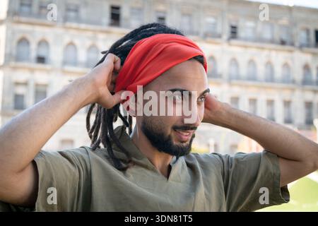 Junger Mann mit einem orangen Bandana auf dem Kopf, der seine Dreadlocks in einer städtischen Umgebung justiert Stockfoto