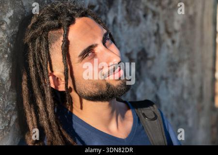 Junger Mann mit Dreadlocks, die lächeln und die Wärme des Sonnenlichts im Gesicht spüren Stockfoto