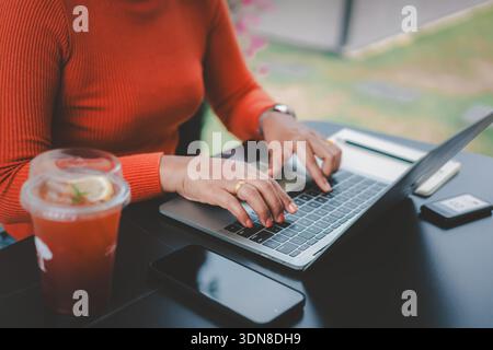 Nahaufnahme einer asiatischen Frau, die in einem Café im Freien auf der Laptop-Tastatur schreibt. Freiberuflicher Mitarbeiter, der online mit Smartphone und Zitronentee arbeitet, digitale Nomade l Stockfoto