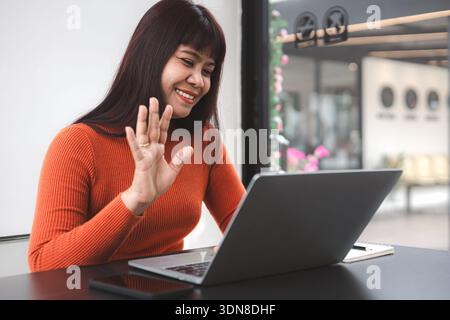 Glückliche asiatische Frau, die während eines Videoanrufs auf dem Laptop im Café Hand winkt. Lächelnde Geschäftsfrau, die Remote arbeitet, Online-Meetings, Lifestyle digitaler Nomaden und t Stockfoto