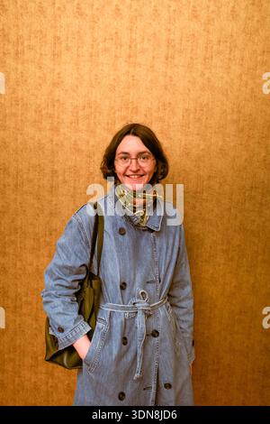 Eine junge Frau im Jeansmantel mit Olivenbeutel steht vor einer strukturierten braunen Wand und lächelt warm. Belgien Stockfoto