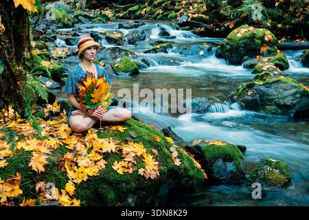 Frau im Wald, die an einem Bach sitzt und Herbstblätter mit hellen Farben hält. Washington State, USA Stockfoto