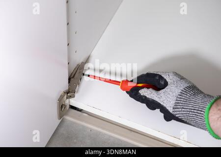 Nahaufnahme der Hand eines Arbeiters in einem grau-schwarzen Schutzhandschuh, der ein metallisches Möbelscharnier an einem weißen Schrank mit einem rot-gelben Schraubendreher justiert Stockfoto