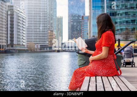 Eine schwangere Mutter sitzt auf einer Bank am Fluss in einer Stadt und liest ein Buch Stockfoto