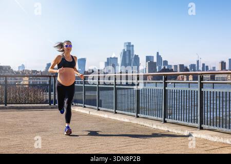 Eine sportliche, schwangere Frau in Sportbekleidung, die an einem sonnigen Tag vor einer Skyline der Stadt zum Sport läuft Stockfoto