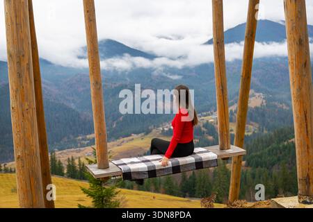 Frau, die sich auf der Schaukel mit Blick auf das Mountain Valley erholt. Frau in rotem Pullover sitzt auf einer Holzschaukel und genießt einen Panoramablick auf die Berge mit nebeligen Wolken und Stockfoto