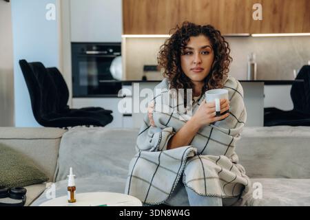 Der weiße Becher liegt in der Hand. Kranke Frau, die sich zu Hause unwohl fühlt, ihre Temperatur überprüft und Medikamente einnimmt, während sie sich auf einem Sofa ausruht. Stockfoto
