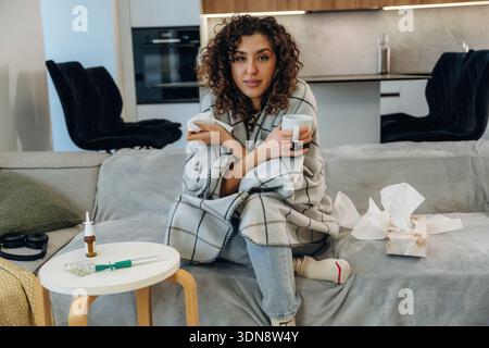 Der weiße Becher liegt in der Hand. Kranke Frau, die sich zu Hause unwohl fühlt, ihre Temperatur überprüft und Medikamente einnimmt, während sie sich auf einem Sofa ausruht. Stockfoto