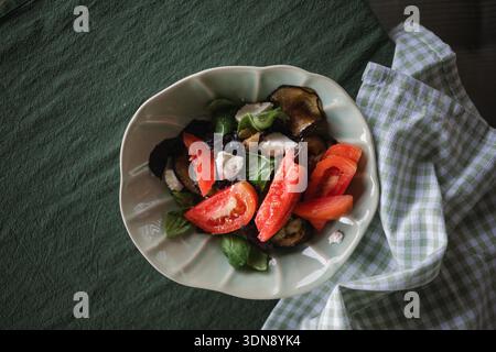 Blick von oben auf eine Schüssel mit frischen Tomaten, gegrillten Auberginen, Fetakäse und mit Olivenöl getränkten Basilikumblättern auf einem Tisch mit Serviette Stockfoto