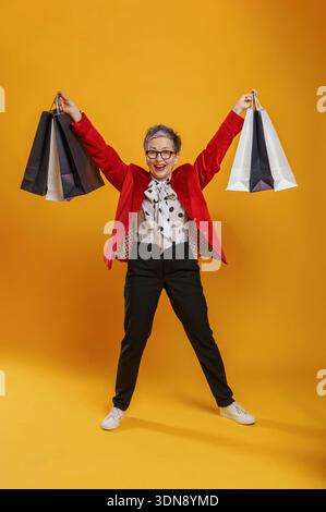 Eine ältere Frau mit grauen Haaren und Brillen hält mit Freude Einkaufstaschen in jeder Hand vor einem hellgelben Hintergrund und zeigt ihre stilvolle Outfit Stockfoto