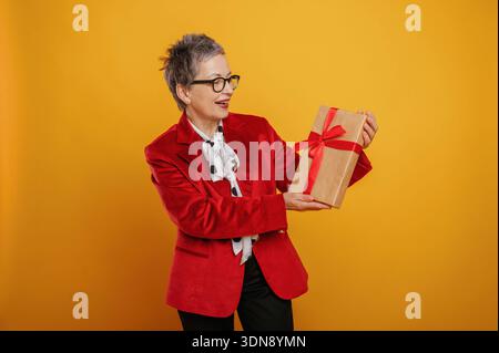 Eine ältere Frau, die einen modischen roten Blazer und einen Schal mit Punktmuster trägt, hält freudig eine Geschenkbox mit rotem Band, die vor einem hellen Schreien steht Stockfoto