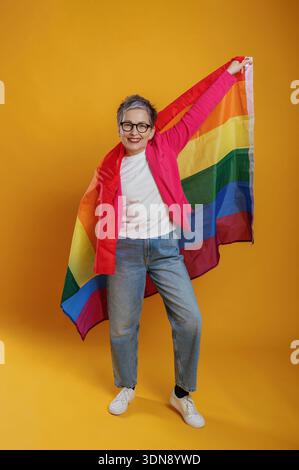 Eine ältere Frau mit grauem Haar, die in einer knalligen rosa Jacke gekleidet ist, hält freudig eine Regenbogenfahne, die Vielfalt und Integration vor einem leuchtenden Geschrei feiert Stockfoto