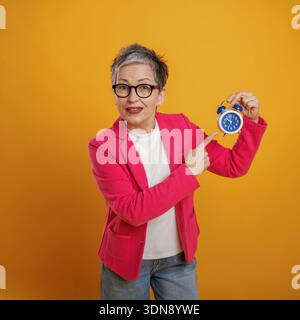 Selbstbewusste Frau mit kurzen grauen Haaren, gekleidet in einem rosafarbenen Blazer, zeigt auf einen blauen Wecker und betont die Bedeutung von Zeitmanagement und p Stockfoto