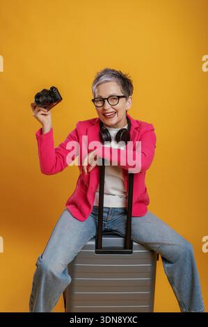 Fröhliche Frau mit kurzen Haaren in einem rosa Blazer hält eine Kamera, während sie auf einem silbernen Koffer sitzt und Begeisterung für Reisen und Werbung ausstrahlt Stockfoto