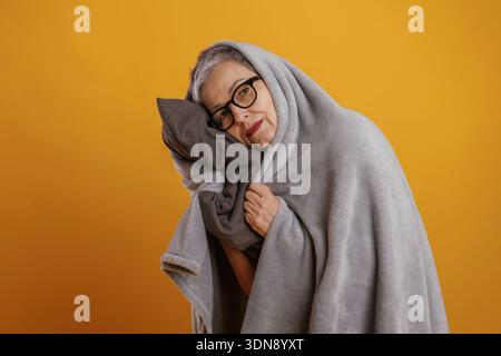 Eine ältere Frau mit kurzen grauen Haaren, die in eine gemütliche graue Decke gewickelt ist, lächelt sanft und umhüllt ein weiches Kissen vor einem leuchtend gelben Rücken Stockfoto